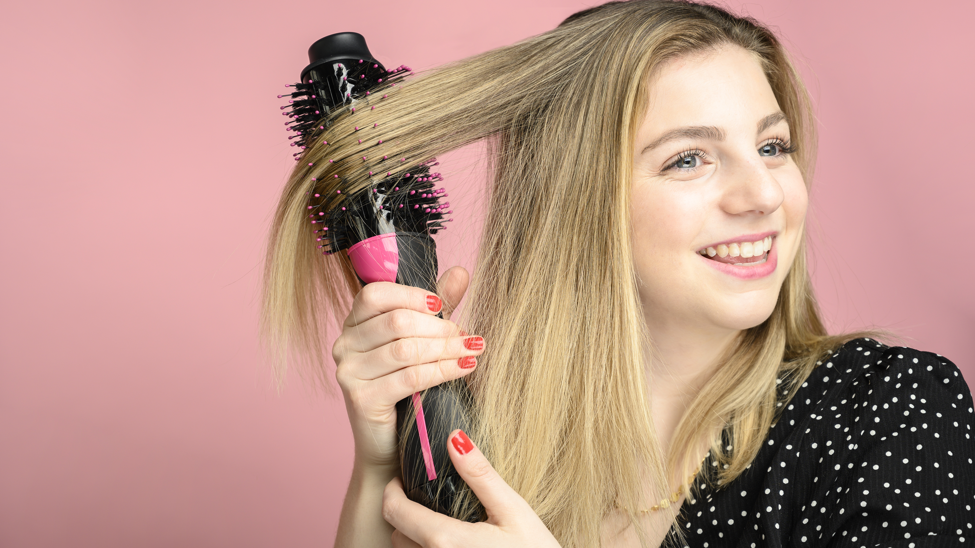 A woman using a hair dryer brush on her blond hair.