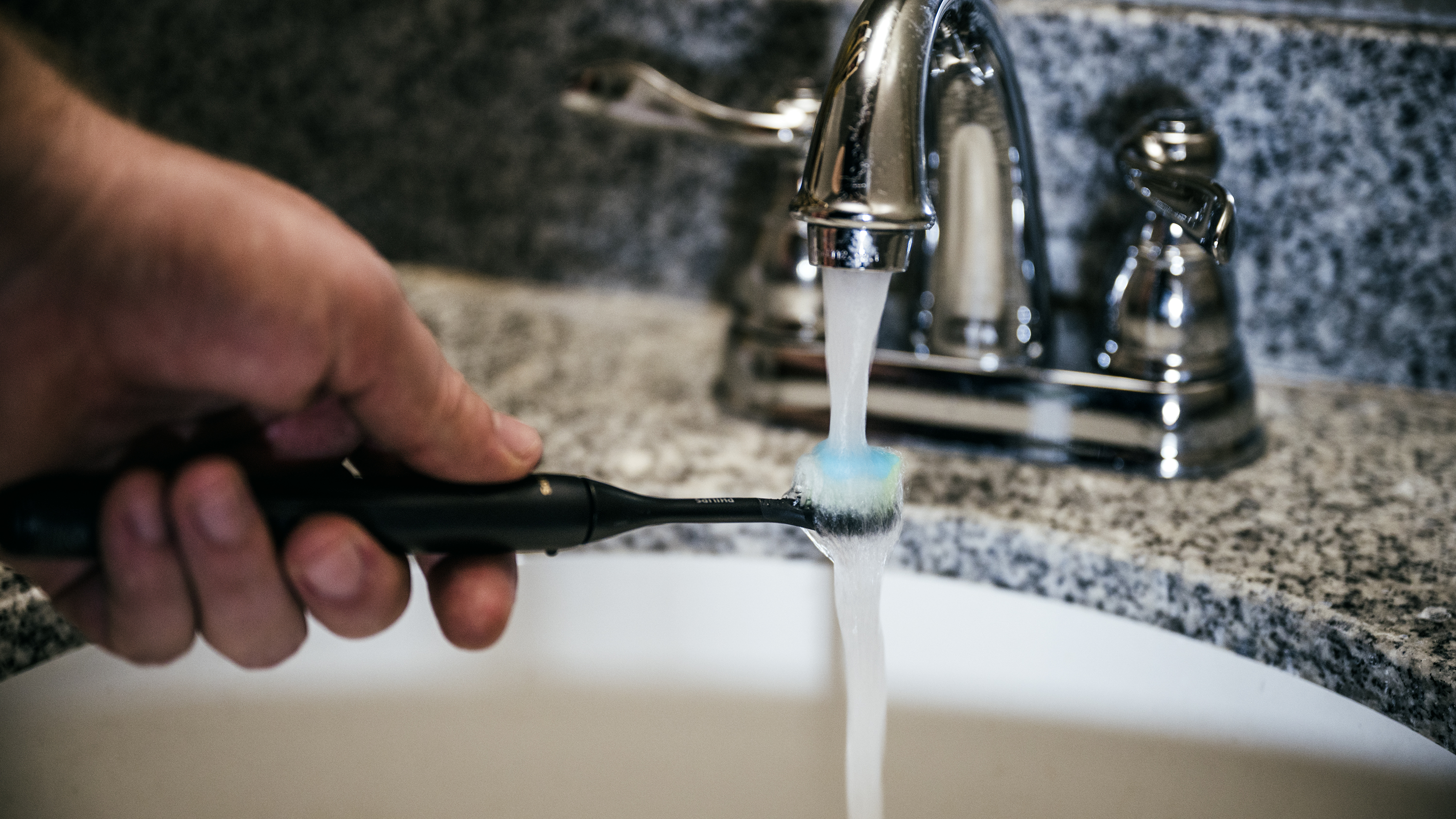 Um homem enxaguar a cabeça de uma escova de dentes elétrica preta sob uma torneira de banheiro
