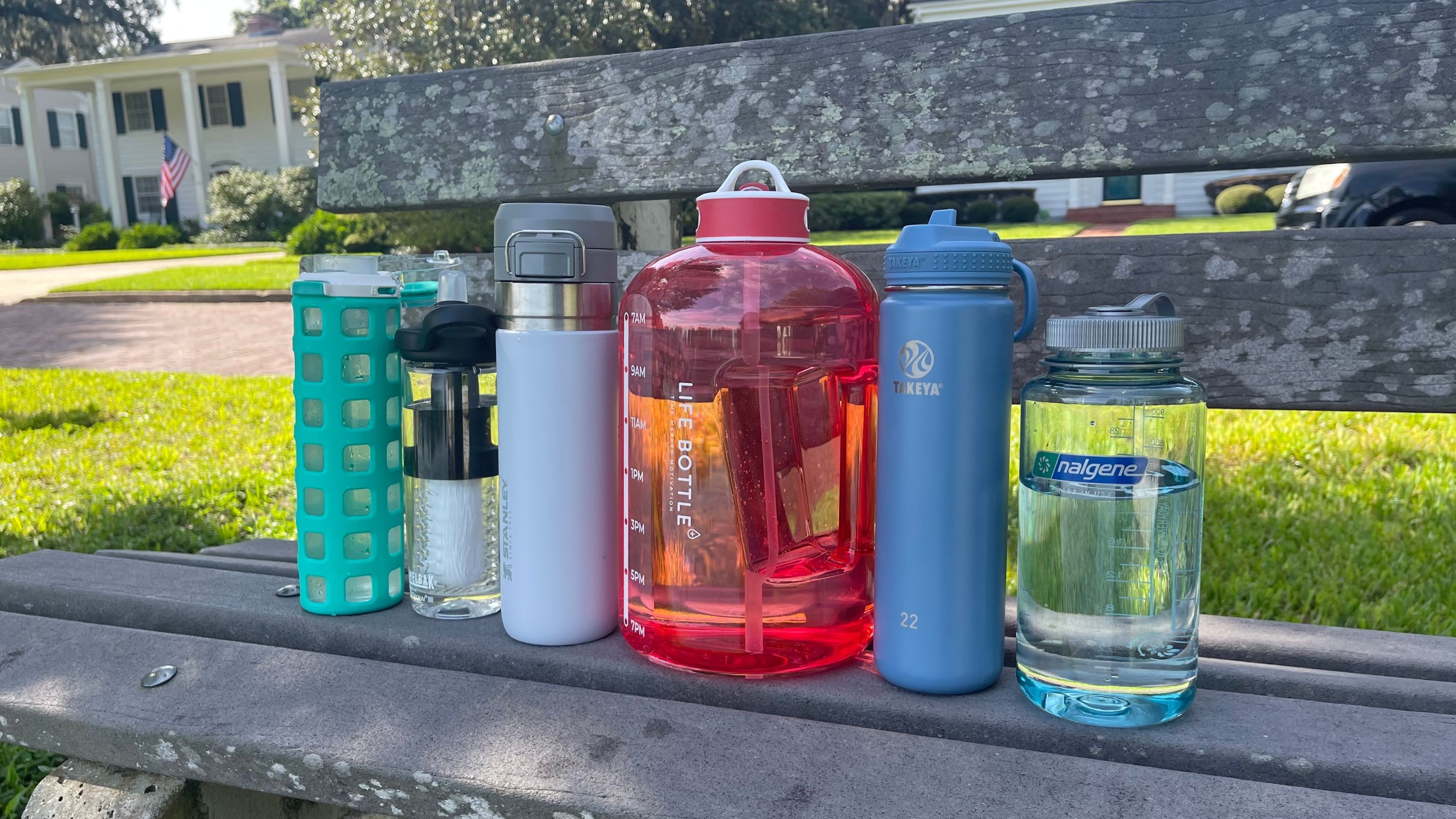 Six reusable water bottles sit on a park bench with a brick road, grass, and homes in the background