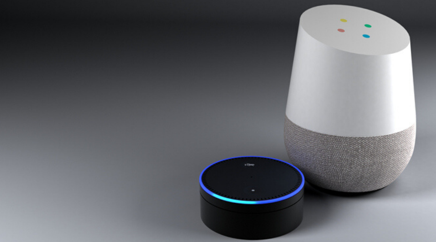 A Google Home speaker and an Amazon Echo Dot speaker sit next to each other.