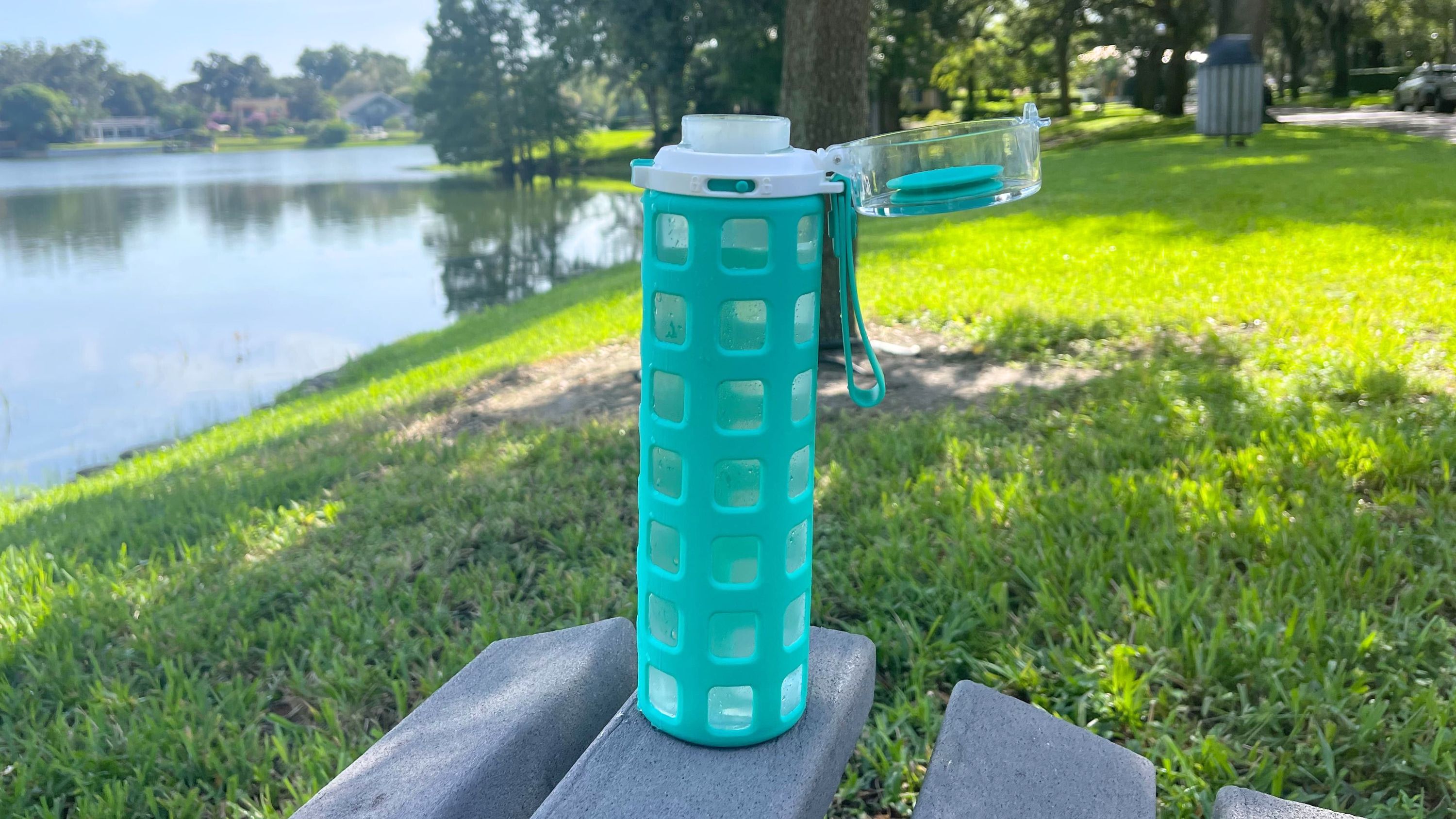 The Ello glass water bottle with green silicone sleeve full of water sitting on a park bench with a lake in the background