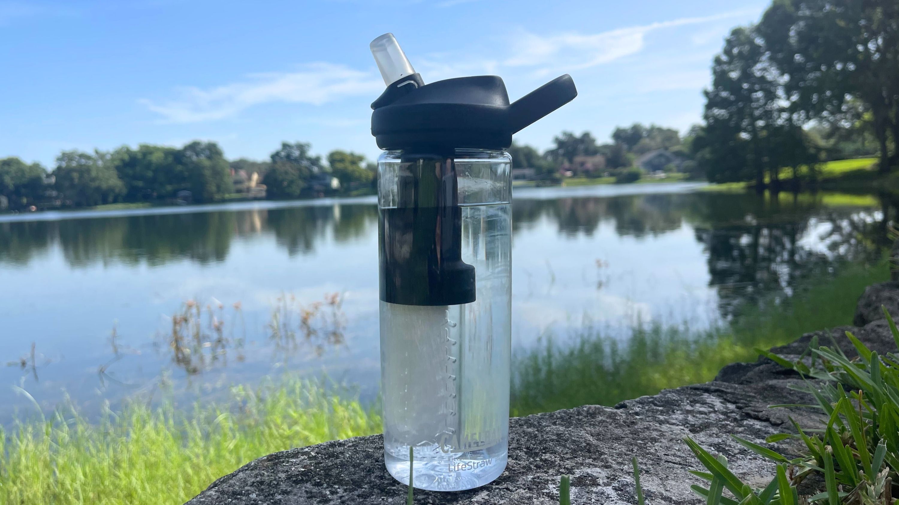 The CamelBak Eddy+ Water Filter Water Bottle by LifeStraw about half full of water, sitting on a stone ledge with a lake in the background