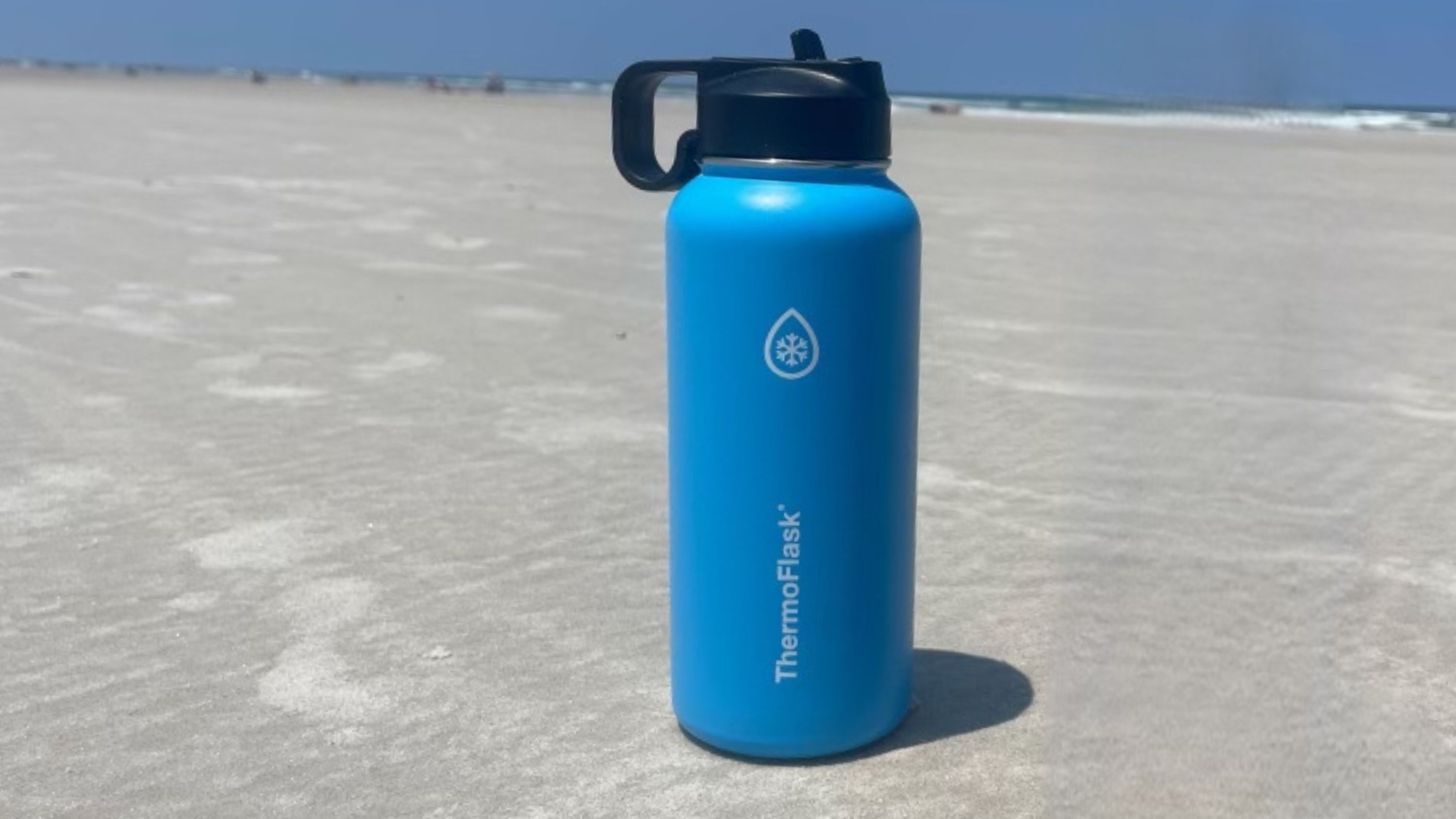 A blue stainless steel water bottle with black lid by ThermoFlask sits on the sand with the ocean in the background